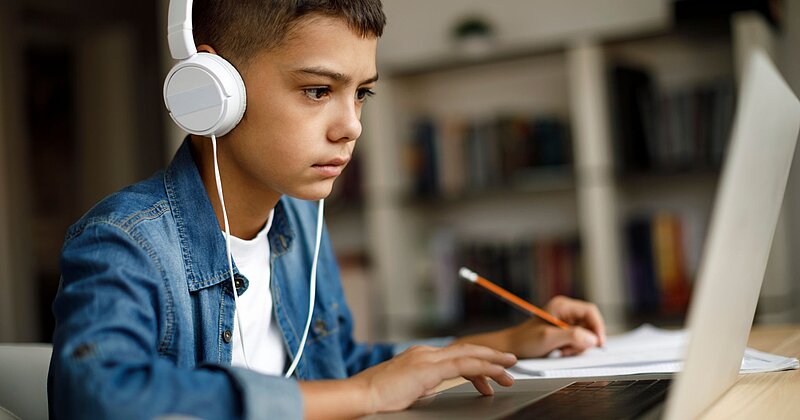 Ein Junge mit Kopfhörern sitzt vor einem Laptop und macht sich auf einem Papierblock Notizen